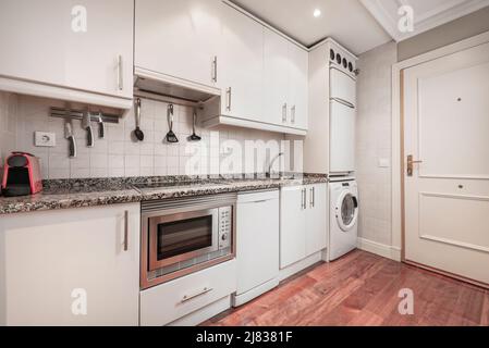 Long apartment kitchen with white cabinets, pink granite countertops, built-in appliances, and brownwood parquet flooring Stock Photo