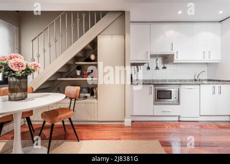 Long apartment kitchen with white cabinets, pink granite countertops, built-in appliances, brown hardwood parquet floors, round dining table and stair Stock Photo