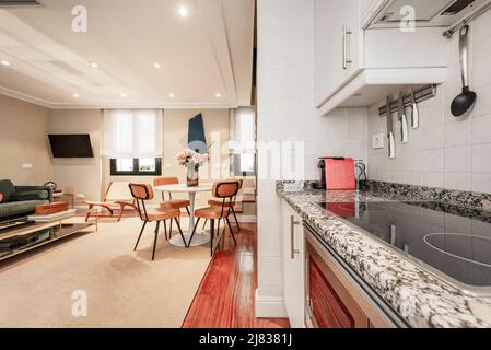 Apartment kitchen with white cabinets, pink granite countertops, built-in appliances, brown hardwood parquet floors, round dining table, and living ro Stock Photo