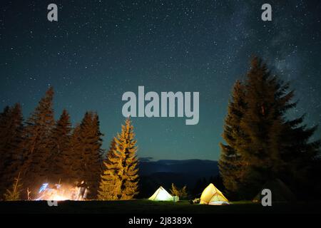 Bright illuminated tourist tents near glowing bonfire on camping site in dark mountain woods under night sky with sparkling stars. Active lifestyle an Stock Photo