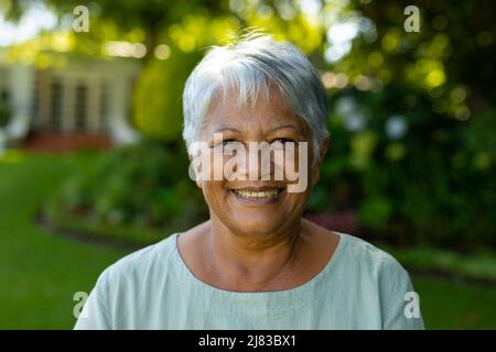 Close-up portrait of smiling biracial senior woman with short gray hair against trees in park Stock Photo