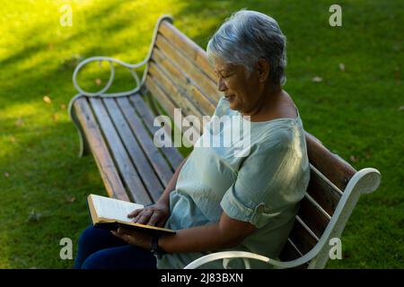 Side view of smiling biracial senior woman with short hair reading book on bench in park Stock Photo