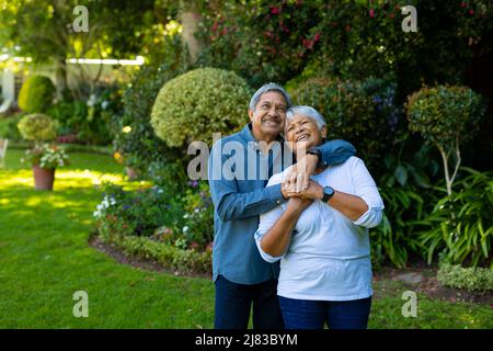Smiling biracial senior couple embracing and looking up while standing against plants in park Stock Photo