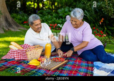 Biracial smiling senior couple with juice on blanket eating strawberries while sitting in park Stock Photo