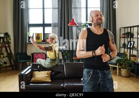 Handsome Caucasian man with tattoo on shoulder wearing wireless headphones enjoying music, his wife taking selfies on background Stock Photo