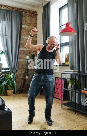 Vertical full body shot of mature Caucasian man wearing jeans and sleeveless shirt enjoying music in wireless headphones and dancing in living room Stock Photo