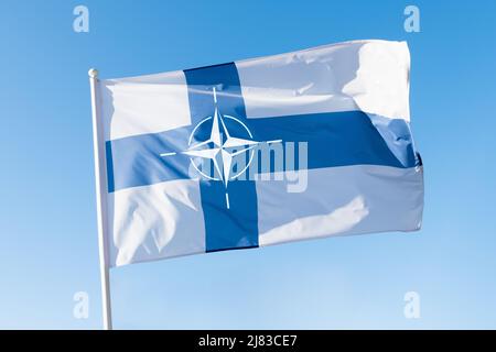 Finland joining NATO concept. Finnish flag with NATO (North Atlantic Treaty Organization) emblem on blue sky background. Stock Photo