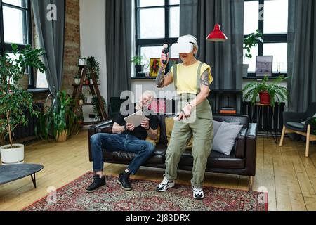 Mature Caucasian man sitting on couch holding digital tablet, his girlfriend enjoying playing video game using VR headset and controllers Stock Photo