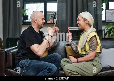 Side view shot of modern mature man and woman in love sitting on couch in living room communicating using sign language Stock Photo