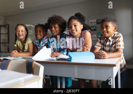 Happy multiracial elementary school students sitting at desk in classroom Stock Photo
