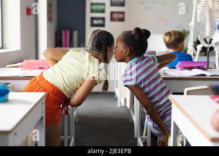 African american elementary girl whispering to caucasian female classmate at desk in classroom Stock Photo