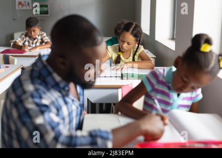 African american young male teacher assisting african american elementary girl studying at desk Stock Photo