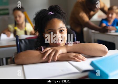 Portrait of smiling biracial elementary schoolgirl sitting on wheelchair at desk in classroom Stock Photo