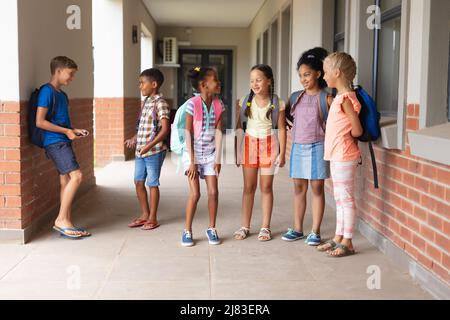 Full length of multiracial elementary school students talking while standing in school corridor Stock Photo