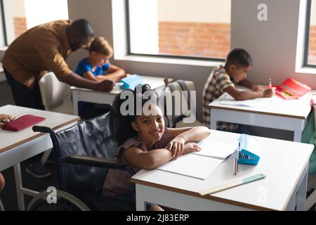 Portrait of smiling biracial elementary schoolgirl sitting on wheelchair at desk in classroom Stock Photo