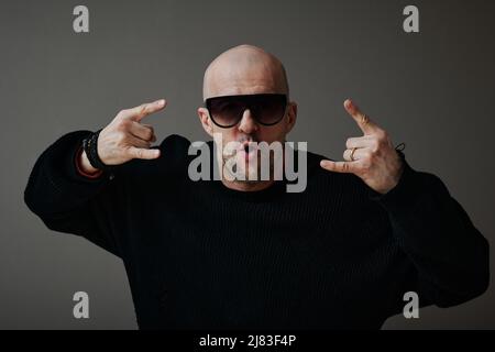 Horizontal waist up studio portrait of cool mature Caucasian man wearing sunglasses and black sweater demonstrating horns sign Stock Photo