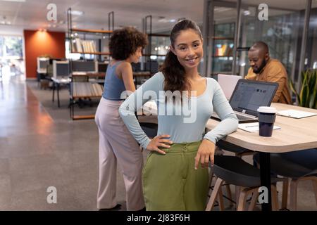 Portrait of smiling young hispanic businesswoman standing by desk against biracial colleagues Stock Photo