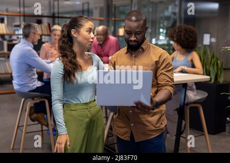 Young biracial businessman and businesswoman discussing over laptop against colleagues at workplace Stock Photo