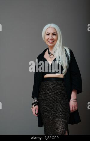 Vertical medium long shot of mature woman wearing fashionable clothes standing against gray wall background, smiling at camera Stock Photo