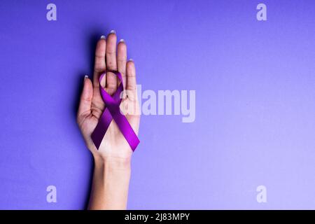 Cropped hand of african american mid adult woman holding purple awareness ribbon on blue background Stock Photo