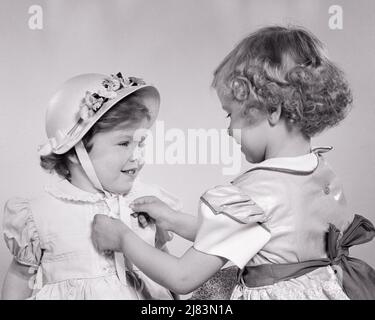 1950s TWO SISTERS GETTING DRESSED OLDER GIRL HELPING YOUNGER TIE HAT RIBBON UNDER HER CHIN - j4812 HAR001 HARS 1 JUVENILE STYLE TEAMWORK STRONG PLEASED JOY SATISFACTION CELEBRATION FEMALES EASTER STUDIO SHOT HEALTHINESS HOME LIFE COPY SPACE FRIENDSHIP HALF-LENGTH CARING FESTIVAL CHIN SIBLINGS CONFIDENCE SISTERS BONNET B&W DRESSES HAPPINESS CHEERFUL EXCITEMENT EASTER BONNET SIBLING SMILES CONNECTION LITTLE SISTER CONCEPTUAL JOYFUL STYLISH PERSONAL ATTACHMENT TYING AFFECTION COOPERATION EMOTION GROWTH JUVENILES TOGETHERNESS YOUNGER BABY GIRL BIG SISTER BLACK AND WHITE CAUCASIAN ETHNICITY HAR001 Stock Photo