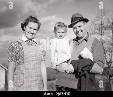 1940s PORTRAIT WORKING MIDDLE CLASS FAMILY MOTHER WEARING APRON FATHER HOLDING TODDLER GIRL AND LUNCH BOX ALL LOOKING AT CAMERA  - j5165 HAR001 HARS THREE HOUSEWIFE ALONE WIFE TODDLER LADY WEARING APRON HUSBAND 3 DAD MOM CLOTHING NOSTALGIC PAIR MOTHERS OLD TIME NOSTALGIA OLD FASHION 1 LUNCHBOX JUVENILE STYLE WELCOME YOUNG ADULT WORKMAN PLEASED FAMILIES JOY LIFESTYLE SATISFACTION FEMALES MARRIED SPOUSE HUSBANDS HOME LIFE COPY SPACE FRIENDSHIP HALF-LENGTH LADIES DAUGHTERS PERSONS CARING MALES CONFIDENCE FATHERS B&W PARTNER EYE CONTACT BLUE COLLAR HOMEMAKER HAPPINESS HOMEMAKERS CHEERFUL AND DADS Stock Photo