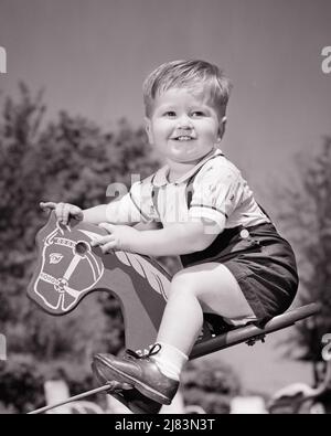 1950s SMILING TODDLER BOY LOOKING AT CAMERA RIDING A STEEL SPRING BOUNCING ROCKING HORSE TOY  - j5413 HAR001 HARS COPY SPACE SEESAW MALES CONFIDENCE B&W HAPPINESS CHEERFUL EXCITEMENT LOW ANGLE A AT PAST SMILES CONCEPTUAL JOYFUL BABY BOY BOUNCING GROWTH JUVENILES SPRINGY BLACK AND WHITE CAUCASIAN ETHNICITY HAR001 OLD FASHIONED Stock Photo