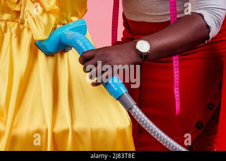 african american tailor dressmaker holding vapor device hot steam generator for silk red and yellow dress in dry-cleaning salon, studio pink