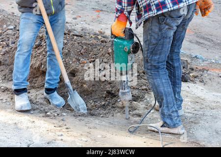 Workers in blue jeans loosen the sandy soil for the construction of a trench at a construction site using an electric jackhammer and shovel. Stock Photo