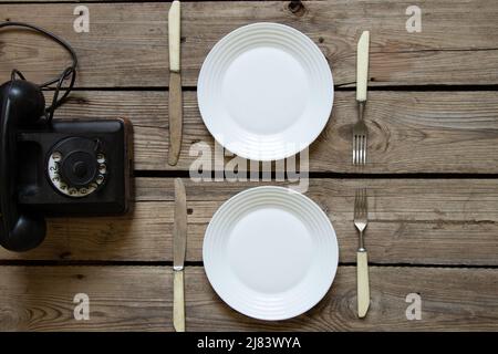 An old retro phone and two plates with knives and forks on a wooden table in the kitchen, ordering food by phone for dinner, a retro phone on the tabl Stock Photo