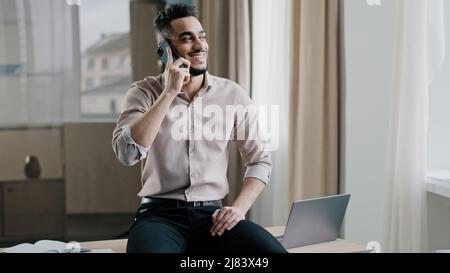 Smiling arab man relaxed male entrepreneur businessman sitting top of table in workspace holding negotiation distant cellphone call with client Stock Photo