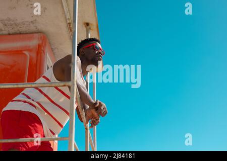 Low angle view of african american young man in sunglasses standing on lifeguard hut against sky Stock Photo