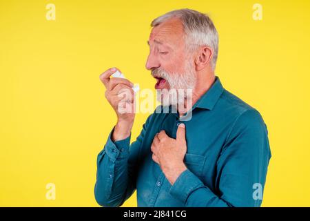 Senior man using an asthma inhaler in studio yellow background Stock Photo