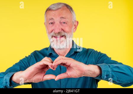 Happy senior man showing hand heart gesture and smiling, take care of the health studio eyllow background Stock Photo