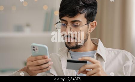 Close-up bearded arabic business man in glasses with credit card and phone makes purchase online shopping enters bank number online transaction uses Stock Photo