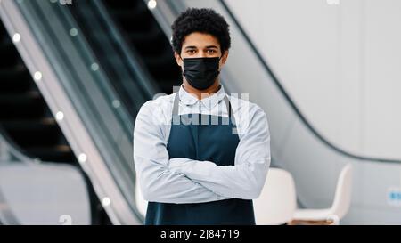 African american man guy restaurant cafe male worker in medical mask looking camera posing crossing arms waiter salesman wearing apron standing at Stock Photo