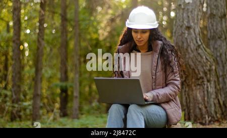 Millennial woman young experienced specialist forestry engineer environmentalist technician in hardhat checking trees entering data into laptop taking Stock Photo