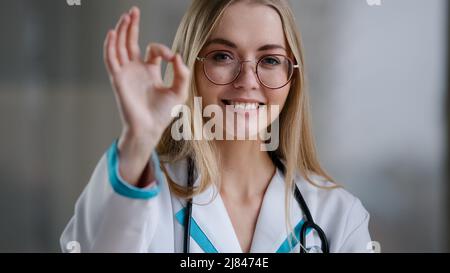 Portrait caucasian happy successful doctor practitioner specialist therapist nurse medical worker woman in glasses looking at camera shows ok gesture Stock Photo