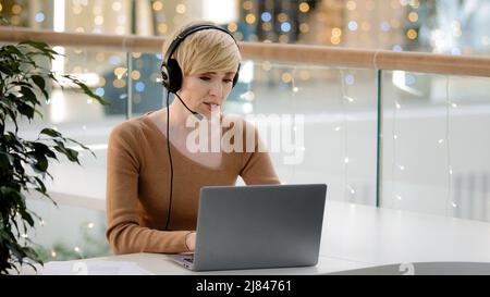 Middle aged adult 40s business woman in headset microphone talking at laptop web camera consultation online distant remote teacher coach helpline Stock Photo