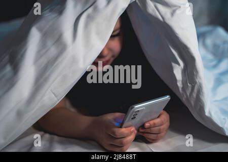 cute little girl hides under blanket with smartphone at night when everyone is asleep. child is smiling and having fun watching cartoons. Children. Blurred Stock Photo