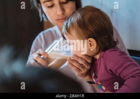 Mother teaching baby girl to drink from glass. Woman helping child drinking milk. Baby care. Stock Photo