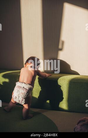 Back view baby girl standing on a sofa in the living room only in a diaper. Happy family leisure. Child development. Family care. Baby development. Stock Photo