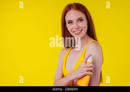 beautiful edheaded ginger woman applying spf sun rays protetor on shoulders arms and neck wearing stylish bikini in studio yellow background Stock Photo
