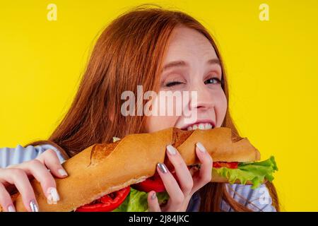 hungry redhaired ginger woman holding huge sandwich with pepper and lettuce salad between bun in studio yellow background Stock Photo