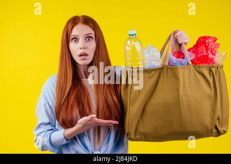 redhead ginger girl with consternation on her face pointing finger copyspase with bad full of non-eco plastic trash in studio yellow background Stock Photo