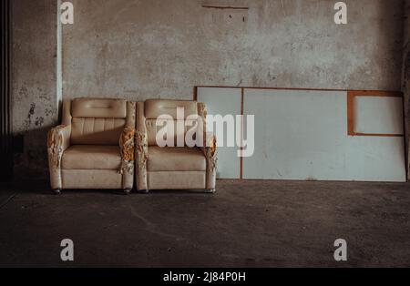 Two old white upholstered armchairs and white planks in front of cement wall in of an abandoned factory site. Copy space, Selective Focus. Stock Photo