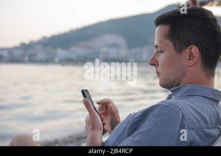 A young man is using his mobile phone while sitting at the beach near the sea. Stock Photo
