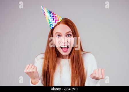 amazed and surprised redhaired ginger woman wearing birthday cap horn holding many gift boxes in studio white background Stock Photo