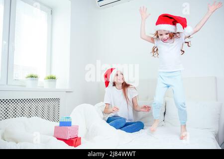 beautiful redhaired ginger woman with santa claus hat and happy little girl in pajamas jumping on bed with white pillow and blanket next to gifts Stock Photo