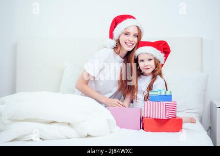 beautiful redhaired ginger woman with santa claus hat and happy little girl in pajamas sitting on bed with white pillow and blanket next to gifts Stock Photo